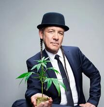 Steve DeAngelo wearing a suit and a hat, holding a small marijuana plant in his palm. 