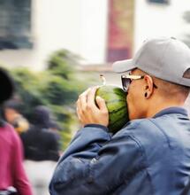 a street scene showing a younger man wearing a grey hat and blue jacket, photographed in profile, he is holding a watermelon bong with in hands and inhaling while moving between other people. 
