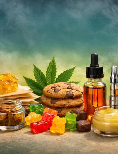 Horizontal image showing an assortment of non-smokable cannabis products arranged on a table, including a bottle of THC syrup with a spoon, golden shatter and wax concentrates, cannabis-infused gummies and cookies, a tincture dropper bottle, vape cartridge, transdermal patch, infused topical cream, capsules, and a cannabis leaf in the background.
