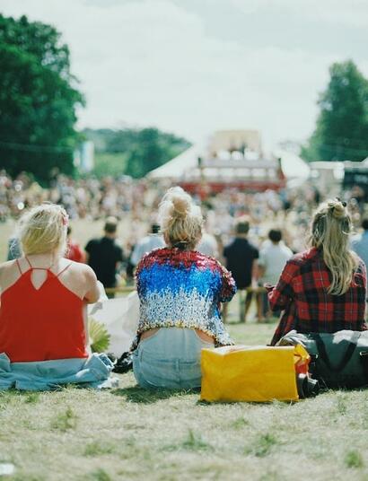 Festival attendees relax on a grassy field under the summer sun while watching performances from a distance at an outdoor music festival.