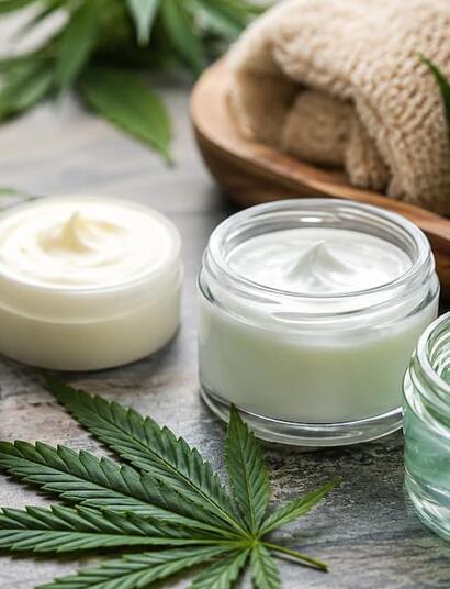 Cannabis topical products including balms, creams, and cooling gels displayed beside cannabis leaves in a spa-style wellness setting.