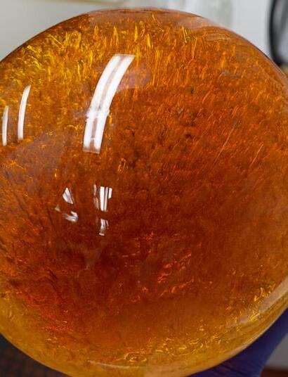 A person wearing a purple glove holds a large spherical glass container filled with a deep amber, resin-like substance. The sphere’s surface reflects overhead lighting, and intricate golden and reddish patterns swirl inside, with lab equipment and a fire extinguisher visible in the background.