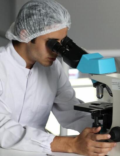 Laboratory technician wearing a white lab coat and hair cover examines a sample through a microscope in a clinical cannabis research setting.