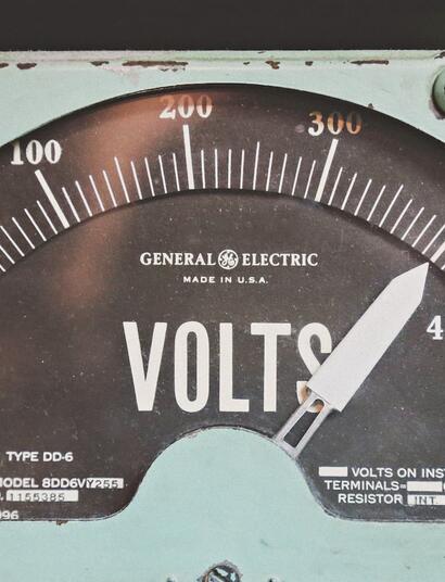 Un voltímetro analógico de estilo industrial con estética retro. La aguja marca un nivel de tensión elevado cerca de la zona roja, sirviendo como metáfora visual de las ráfagas de hiperexcitabilidad eléctrica en el cerebro que el CBD y el CBDV ayudan a regular según el estudio de Iannotti y Di Marzo.