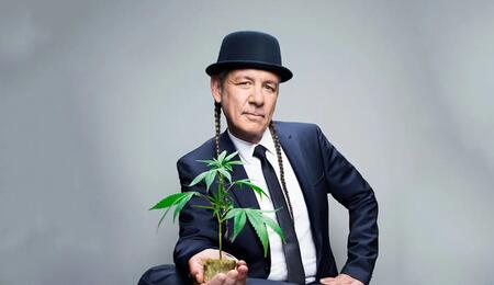 Steve DeAngelo wearing a suit and a hat, holding a small marijuana plant in his palm. 