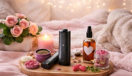 Romantic cannabis wellness flat lay featuring premium vaporizers, heart-shaped edibles, infused massage oil, candles, and pink roses arranged on a wooden tray atop a soft blanket, creating an intimate Valentine’s Day atmosphere.