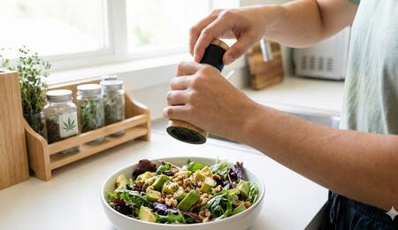 Una persona preparando una ensalada saludable y moliendo pimienta negra, ilustrando el concepto de "cannabinoide dietético" del betacariofileno, un potente antiinflamatorio natural que activa el receptor CB2.