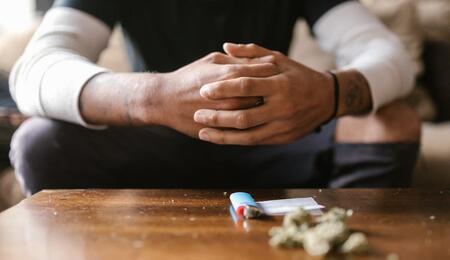 Person sitting with hands clasped at a table, cannabis buds and a lighter placed in the foreground, symbolizing a cannabis tolerance break and mindful pause from consumption.