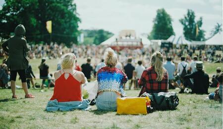 Festival attendees relax on a grassy field under the summer sun while watching performances from a distance at an outdoor music festival.