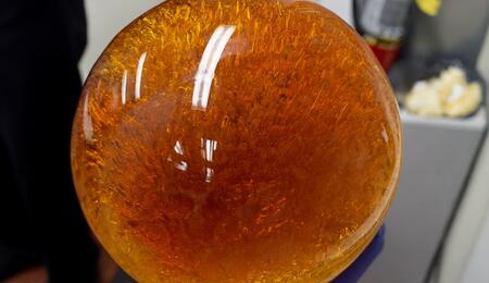 A person wearing a purple glove holds a large spherical glass container filled with a deep amber, resin-like substance. The sphere’s surface reflects overhead lighting, and intricate golden and reddish patterns swirl inside, with lab equipment and a fire extinguisher visible in the background.