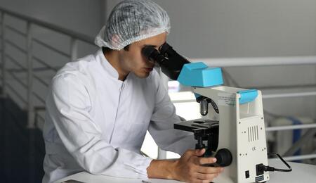 Laboratory technician wearing a white lab coat and hair cover examines a sample through a microscope in a clinical cannabis research setting.