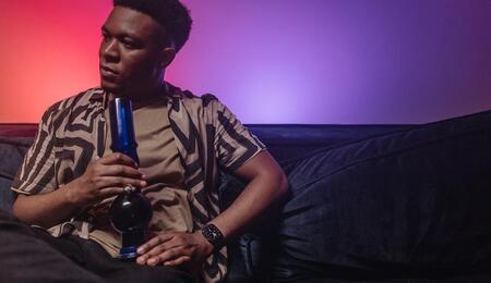A young black man sitting on a sofa and holding a bong for smoking weed in his hands.