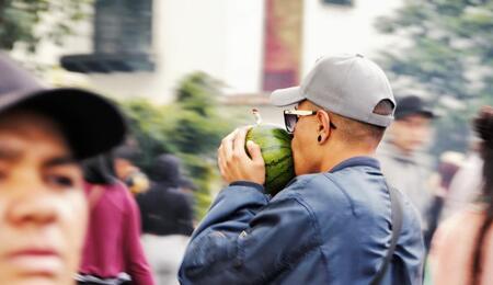a street scene showing a younger man wearing a grey hat and blue jacket, photographed in profile, he is holding a watermelon bong with in hands and inhaling while moving between other people. 