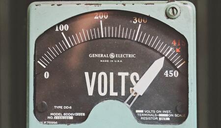 Un voltímetro analógico de estilo industrial con estética retro. La aguja marca un nivel de tensión elevado cerca de la zona roja, sirviendo como metáfora visual de las ráfagas de hiperexcitabilidad eléctrica en el cerebro que el CBD y el CBDV ayudan a regular según el estudio de Iannotti y Di Marzo.