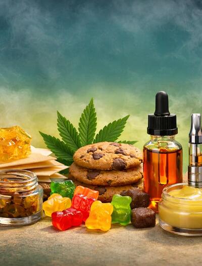Horizontal image showing an assortment of non-smokable cannabis products arranged on a table, including a bottle of THC syrup with a spoon, golden shatter and wax concentrates, cannabis-infused gummies and cookies, a tincture dropper bottle, vape cartridge, transdermal patch, infused topical cream, capsules, and a cannabis leaf in the background.