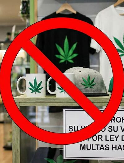Interior de un grow shop con una señal de prohibido roja sobre productos de merchandising (camisetas, tazas, gorras) con la hoja de marihuana. Un cartel advierte de la prohibición de venta y regalo por la Ley de Galicia con multas de hasta 600.000€.