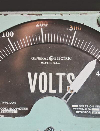 Un voltímetro analógico de estilo industrial con estética retro. La aguja marca un nivel de tensión elevado cerca de la zona roja, sirviendo como metáfora visual de las ráfagas de hiperexcitabilidad eléctrica en el cerebro que el CBD y el CBDV ayudan a regular según el estudio de Iannotti y Di Marzo.