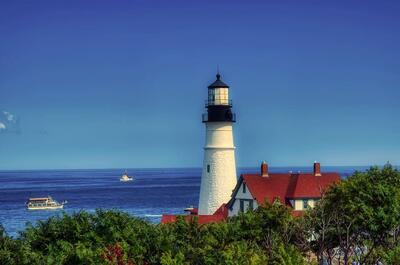 Historic lighthouse overlooking the rocky Maine coastline under a clear blue sky, surrounded by greenery and calm Atlantic waters.