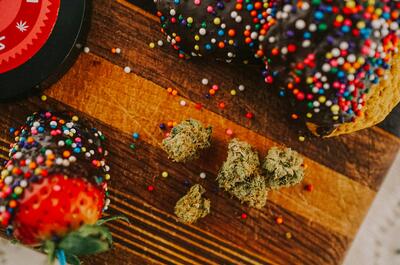 A close-up of high-THC cannabis buds on a wooden surface, surrounded by a chocolate-covered strawberry and dessert sprinkled with colorful nonpareils. A portion of a red container with a cannabis leaf symbol is visible in the corner.