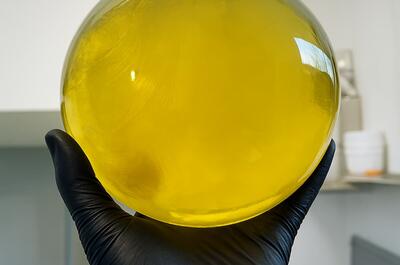 A gloved hand holds a large, round glass vessel filled with a bright, translucent yellow liquid. Light reflections and subtle swirling textures appear within the liquid, while the background shows a blurred laboratory setting.