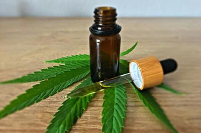 an image of an open brown bottle with a dropper containing cannabis oil, laid on a table with marijuana fan leaf.