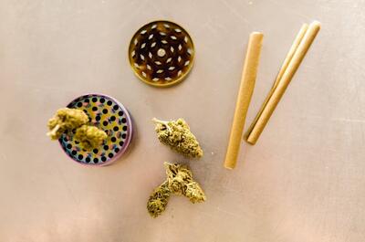 Overhead view of cannabis flower buds, a colorful metal grinder, and several pre-rolled cones arranged on a tabletop.