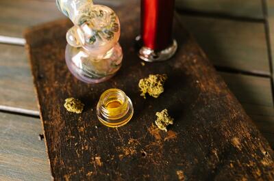 Cannabis concentrate in a small glass container displayed alongside flower buds, a glass rig, and a vaporizer on a rustic wooden surface.