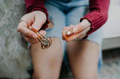 Person holding a glass cannabis pipe and a lighter while seated indoors, preparing for a smoking session.