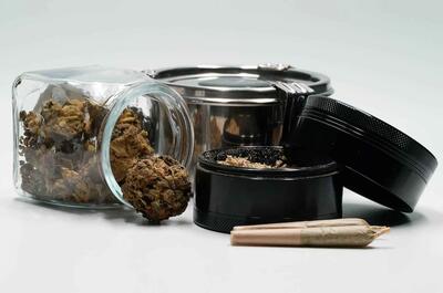 An image showing an open jar with buds of weed put next to a black grinder with weed, another metal container, and two ready-made pre-roll joints.