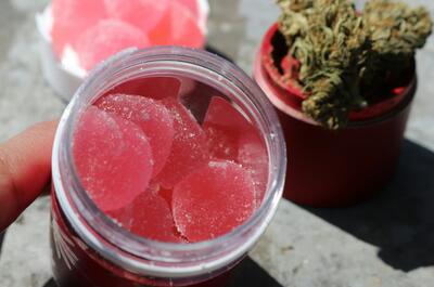 photo showing a jar with mouthwatering watermelon pink gummies and a grinder in the background with puffy buds of weed.