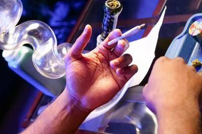 Close-up of a person holding a lit cannabis joint over a glass table, with a glass bong and cannabis buds visible nearby under blue lighting. 